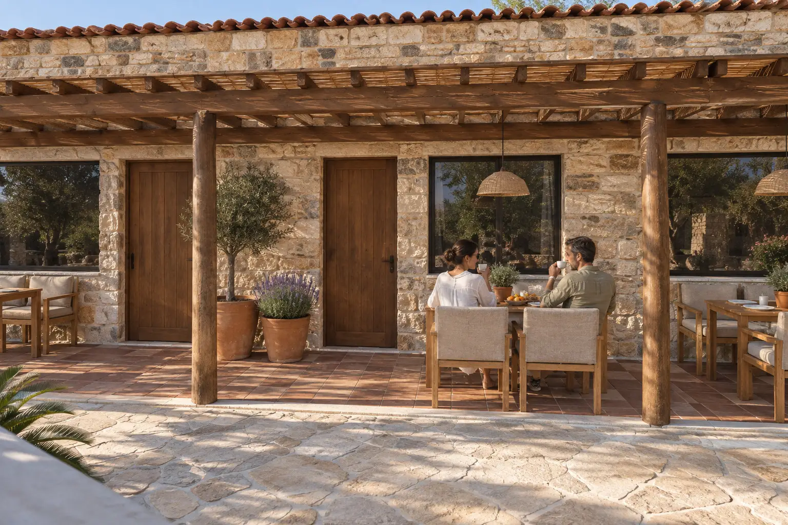 Zwei Personen an einem Esstisch unter einer Holzpergola vor einem Steinhaus mit Glastüren und Topfpflanzen.