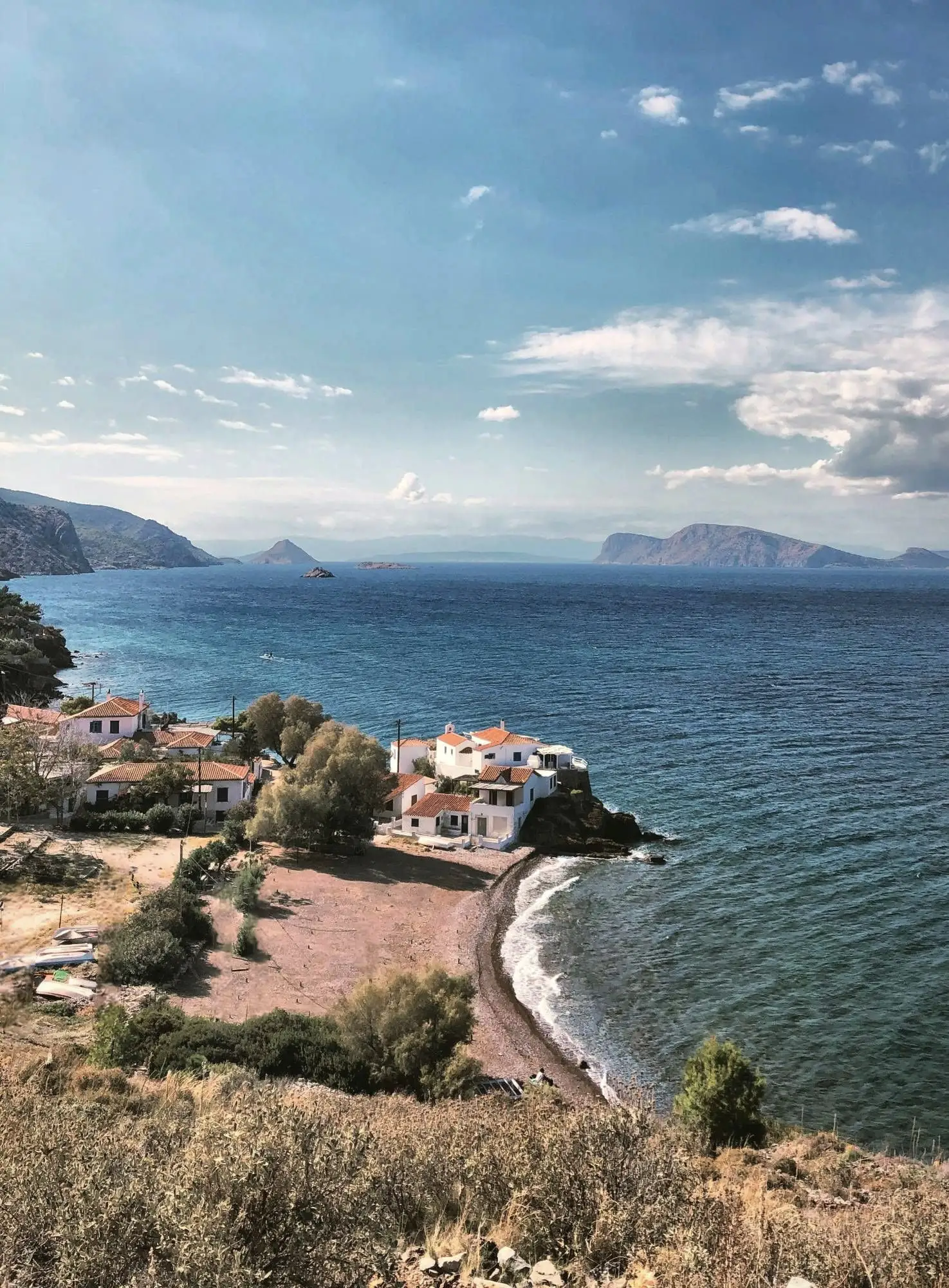 White houses on a rocky point above a small pebble beach, with islands and the Peloponnese across the sea