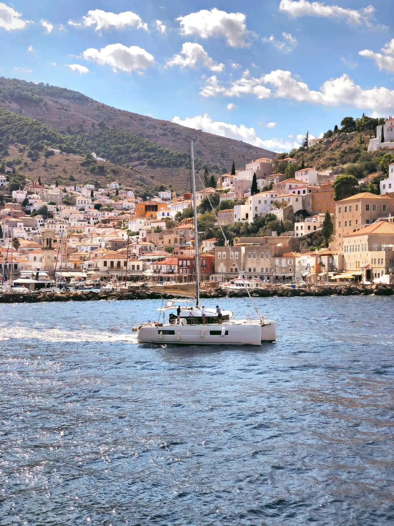 A catamaran crossing in front of Hydra town, its stone houses climbing the hill above the harbour
