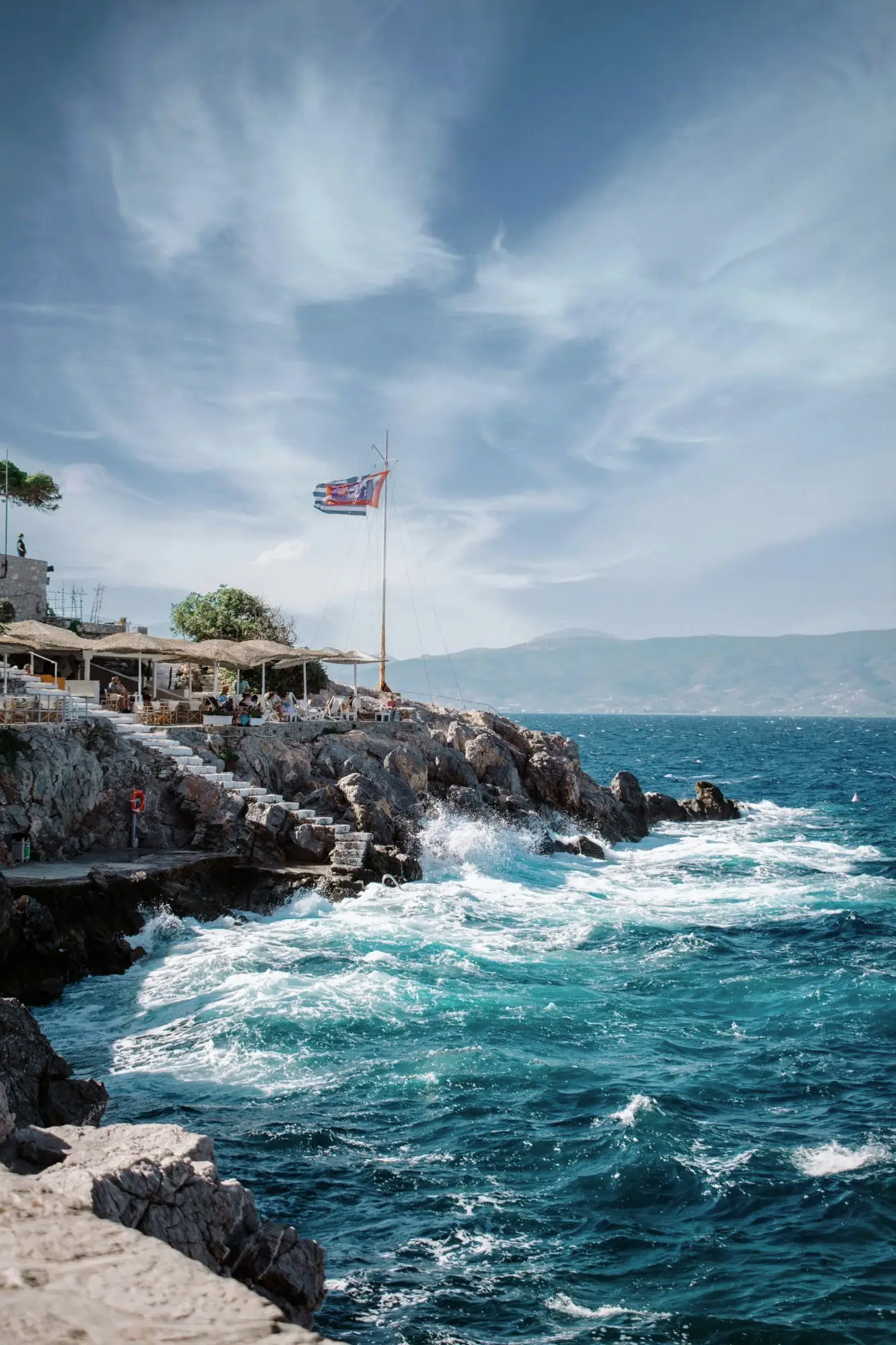 Waves breaking on the swimming rocks at Spilia, with white steps, parasols and a Greek flag above the sea