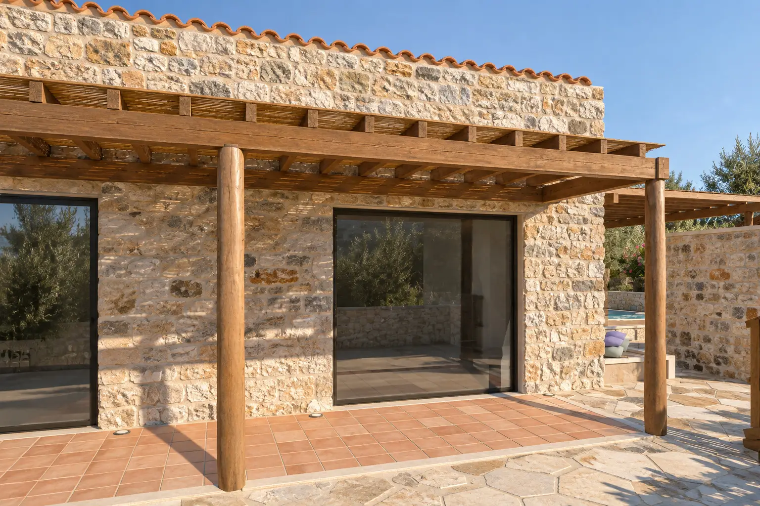 Steinfassade mit breiten Glastüren hinter einer Holzpergola und einer sonnigen Terrasse mit Terrakottafliesen.